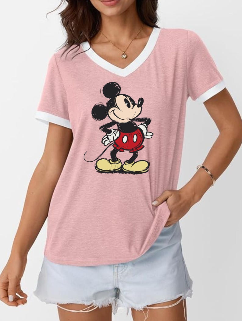 May include: Pink V-neck t-shirt with white trim featuring a cartoon illustration of Mickey Mouse. Mickey is depicted in his classic outfit: red shorts, yellow shoes, and white gloves. The shirt has short sleeves.