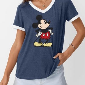 May include: Navy blue V-neck t-shirt with white trim featuring a graphic of Mickey Mouse. The cartoon character is in a classic pose, wearing red shorts, yellow shoes, and white gloves. A casual, comfortable top.