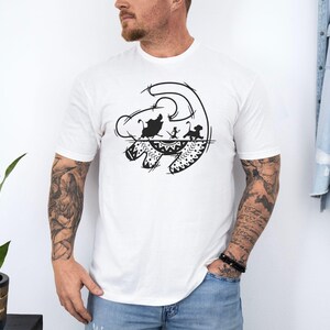 May include: White t-shirt featuring a black graphic design of a lion's face with silhouettes of characters from The Lion King. The design includes a stylized sun and decorative patterns. The shirt is short-sleeved.