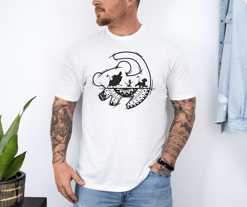 May include: White t-shirt featuring a black graphic design of a lion's face with silhouettes of characters from The Lion King. The design includes a stylized sun and decorative patterns. The shirt is short-sleeved.