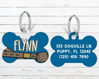 May include: Blue dog bone-shaped pet tag with the name "Flynn" in a bold font and a cartoon belt design. The reverse side displays contact information: address, city, zip code, and phone number.
