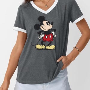 May include: A gray V-neck t-shirt with white trim featuring a graphic of Mickey Mouse. Mickey is depicted with his signature black ears, red shorts, yellow shoes, and white gloves. The shirt is a casual style.