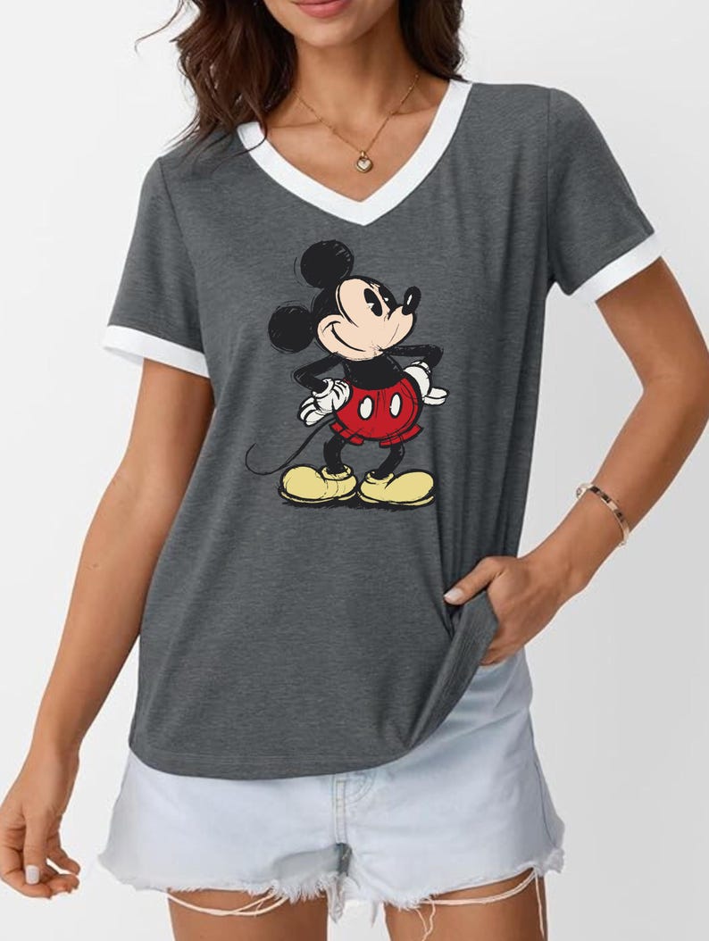 May include: A gray V-neck t-shirt with white trim featuring a graphic of Mickey Mouse. Mickey is depicted with his signature black ears, red shorts, yellow shoes, and white gloves. The shirt is a casual style.