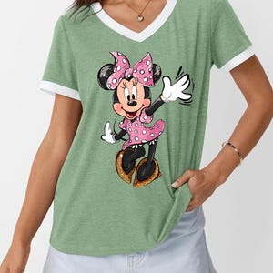 K&ouml;nnte beinhalten: Gr&uuml;nes V-Ausschnitt-T-Shirt mit wei&szlig;em Besatz mit einem Minnie Mouse-Motiv. Minnie tr&auml;gt ein rosa gepunktetes Kleid und eine Schleife mit goldenen Glitzerschuhen. Das Shirt hat einen l&auml;ssigen Stil.