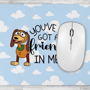 May include: A rectangular mousepad with a light blue sky and white cloud design. The mousepad features a cartoon dog character and the text "YOU'VE GOT A FRIEND IN ME". A white wireless mouse is on the right side of the mousepad.