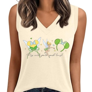 May include: Cream-colored sleeveless V-neck top featuring a graphic of Tinkerbell, a bow, and Minnie Mouse ears. The text "Fly with faith and trust" is written in a script font. The top is paired with blue jeans.