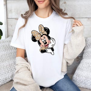 May include: White t-shirt with a Minnie Mouse cartoon graphic. The design shows Minnie Mouse with a polka-dot bow, black ears, and a tan and white outfit. The t-shirt has a classic crew neck.