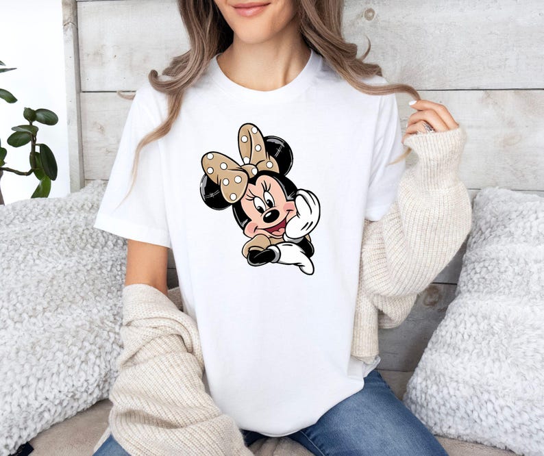 May include: White t-shirt with a Minnie Mouse cartoon graphic. The design shows Minnie Mouse with a polka-dot bow, black ears, and a tan and white outfit. The t-shirt has a classic crew neck.