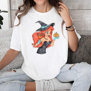 May include: White t-shirt featuring a cartoon witch with red hair, a black witch hat, and a black dress. The witch is holding a pumpkin and a candy apple. The design has a Halloween theme.
