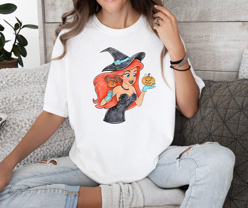 May include: White t-shirt featuring a cartoon witch with red hair, a black witch hat, and a black dress. The witch is holding a pumpkin and a candy apple. The design has a Halloween theme.