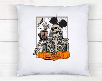 May include: White square pillow featuring a Halloween-themed illustration. A skeleton wearing Mickey Mouse ears holds a coffee cup with a castle design. Orange jack-o'-lanterns sit below the skeleton, with a spooky background.