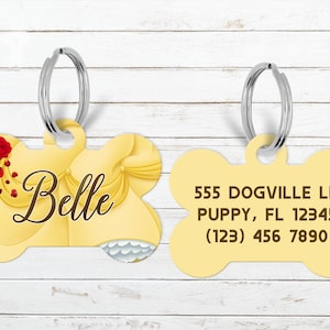 May include: A yellow dog tag in a bone shape with a silver ring. One side features the name "Belle" in script with a rose and dress design. The other side has an address and phone number.