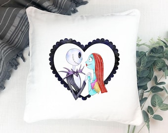 May include: White square pillow featuring a heart-shaped design with Jack Skellington and Sally from The Nightmare Before Christmas. The characters are in a black scalloped border. The pillow is on a white surface.