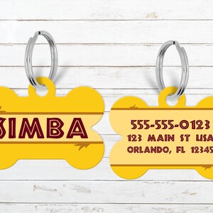 May include: Two yellow bone-shaped pet ID tags with silver key rings. One tag has the name "SIMBA" in maroon lettering. The other tag displays a phone number, address, and city in maroon lettering.