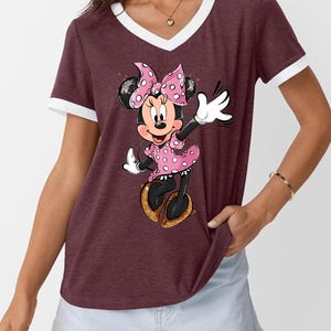 K&ouml;nnte beinhalten: Burgunderfarbenes V-Ausschnitt-T-Shirt mit wei&szlig;em Besatz mit einem Minnie Mouse-Motiv. Minnie tr&auml;gt ein rosa gepunktetes Kleid und eine Schleife mit goldenen Schuhen. Das Shirt ist ein l&auml;ssiger Stil.