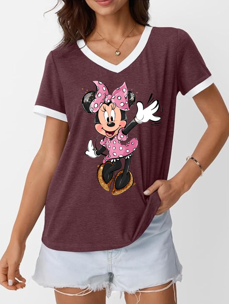 K&ouml;nnte beinhalten: Burgunderfarbenes V-Ausschnitt-T-Shirt mit wei&szlig;em Besatz mit einem Minnie Mouse-Motiv. Minnie tr&auml;gt ein rosa gepunktetes Kleid und eine Schleife mit goldenen Schuhen. Das Shirt ist ein l&auml;ssiger Stil.