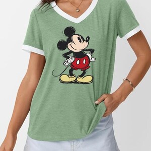 May include: Green V-neck t-shirt with white trim featuring a graphic of Mickey Mouse. The cartoon character is depicted in his classic outfit: red shorts, yellow shoes, and white gloves. The shirt is a casual style.