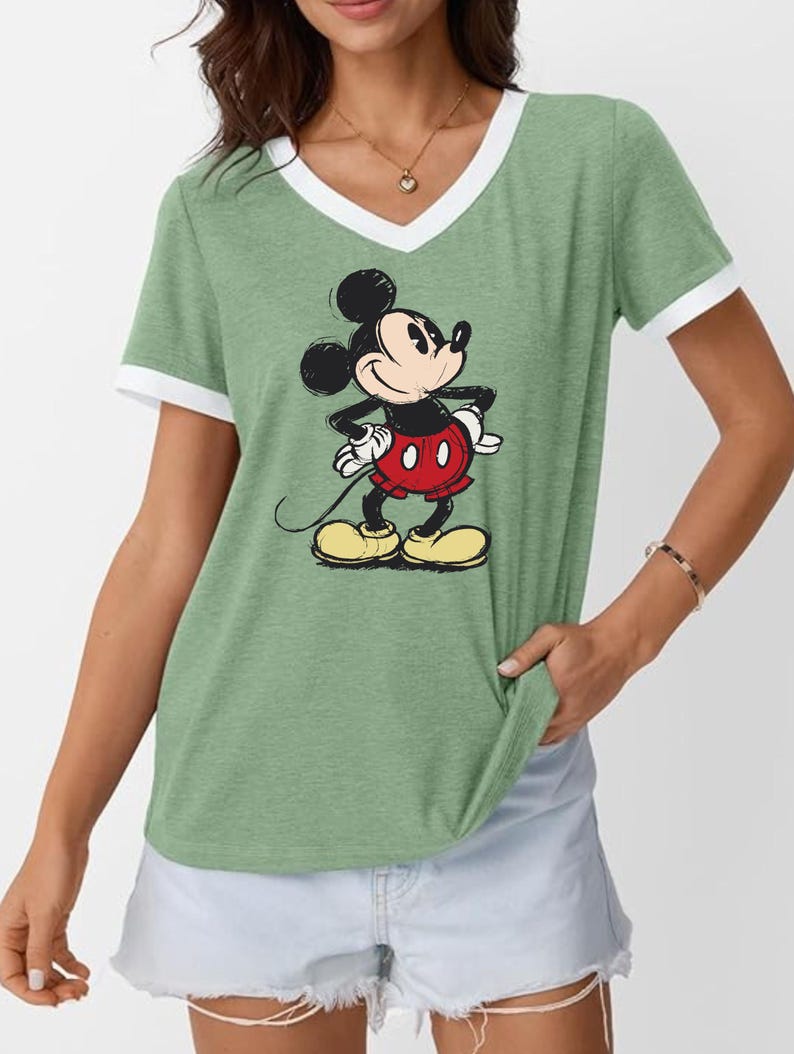 May include: Green V-neck t-shirt with white trim featuring a graphic of Mickey Mouse. The cartoon character is depicted in his classic outfit: red shorts, yellow shoes, and white gloves. The shirt is a casual style.
