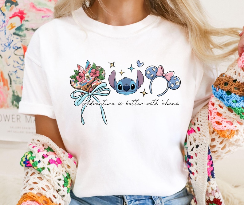 May include: White t-shirt featuring a colorful graphic of Stitch, a ukulele, and Mickey Mouse ears. The design includes the text "Adventure is better with 'ohana" in a script font. The shirt is a casual, everyday wear item.
