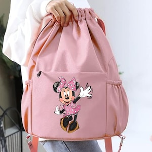 May include: Pink drawstring backpack featuring a Minnie Mouse graphic. The backpack has a front pocket and side pockets. The Minnie Mouse graphic shows Minnie in a pink polka dot dress and bow, waving her hand. The backpack is held by a person.