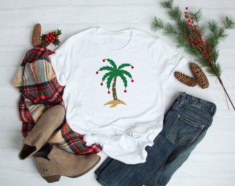 May include: White t-shirt featuring a green palm tree decorated with red ornaments and string lights. The palm tree is on a sandy island. The shirt is paired with blue jeans, a plaid scarf, and brown boots, suggesting a holiday theme.