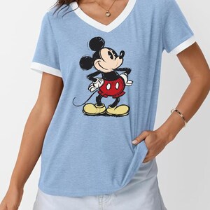 May include: Light blue V-neck t-shirt with white trim and a graphic of Mickey Mouse. The cartoon character is in a classic pose, wearing red shorts and yellow shoes. The shirt is a casual style.