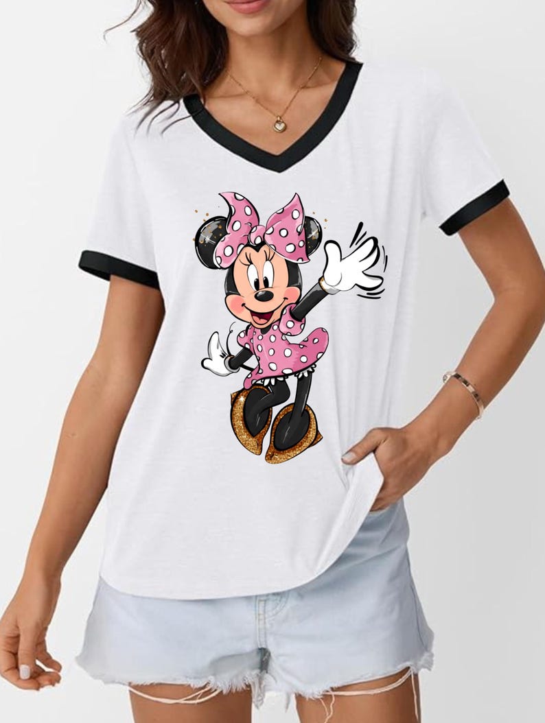 K&ouml;nnte beinhalten: Wei&szlig;es V-Ausschnitt-T-Shirt mit schwarzem Besatz mit einem Cartoon-Minnie-Mouse-Design. Minnie Mouse tr&auml;gt ein rosa gepunktetes Kleid und eine Schleife mit goldenen Glitzerschuhen. Das Shirt ist ein l&auml;ssiger Stil.