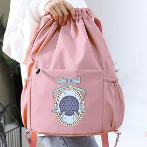 May include: A pink drawstring backpack with a front pocket and side pockets. The backpack features a graphic of a blue and purple sphere with a bow and decorative frame. The straps are a lighter pink.