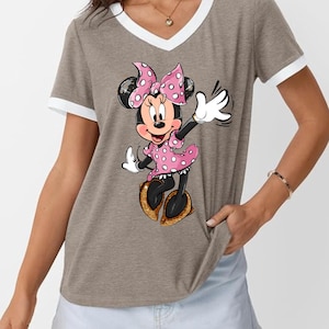 K&ouml;nnte beinhalten: Taupefarbenes V-Ausschnitt-T-Shirt mit wei&szlig;em Besatz an Ausschnitt und &Auml;rmeln. Das Shirt zeigt eine Cartoon-Illustration von Minnie Mouse in einem rosa gepunkteten Kleid und einer Schleife, die mit einem goldenen Glitzerschuh winkt.
