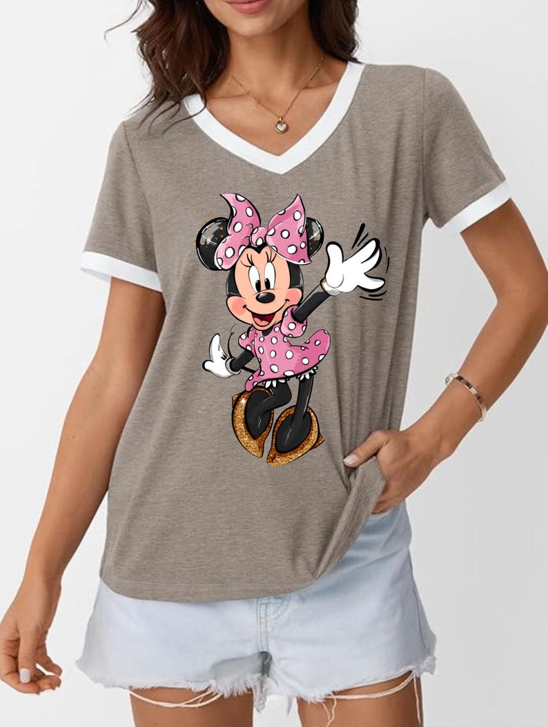 K&ouml;nnte beinhalten: Taupefarbenes V-Ausschnitt-T-Shirt mit wei&szlig;em Besatz an Ausschnitt und &Auml;rmeln. Das Shirt zeigt eine Cartoon-Illustration von Minnie Mouse in einem rosa gepunkteten Kleid und einer Schleife, die mit einem goldenen Glitzerschuh winkt.