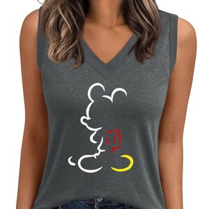 Puede incluir: Una camiseta sin mangas gris con cuello en V y un contorno blanco de Mickey Mouse. El pantal&oacute;n corto del personaje de dibujos animados es amarillo y su pajarita es roja. La camiseta se combina con unos vaqueros azules.