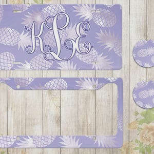 May include: A rectangular license plate, a license plate frame, and two circular car coasters, all featuring a light purple background with a repeating white pineapple pattern and the monogram letters K, L, and E in a decorative script font.
