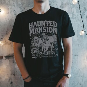 May include: Black t-shirt with a gray graphic design featuring the words "Haunted Mansion" and various spooky illustrations. The design includes text that reads "Foolish Mortals" and "Beware of Hitchhiking Ghosts".