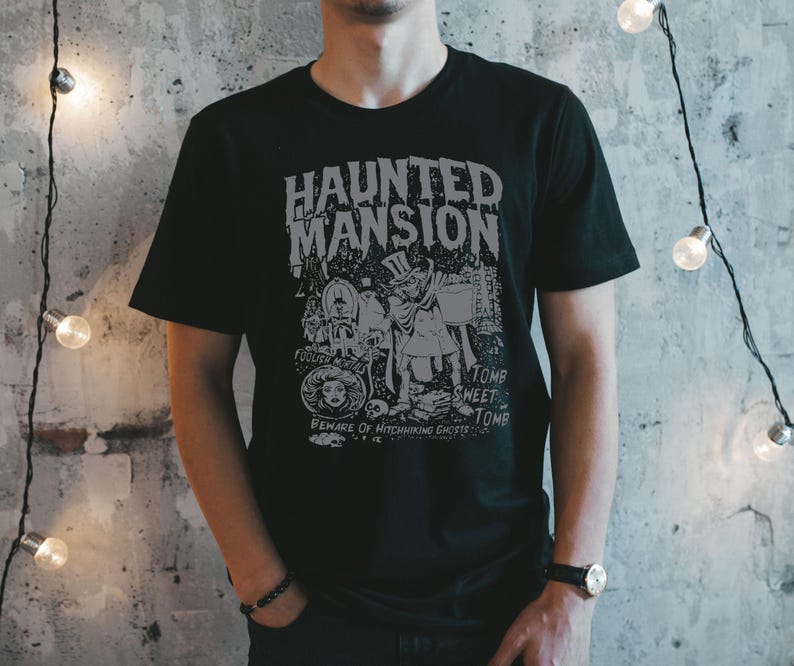 May include: Black t-shirt with a gray graphic design featuring the words "Haunted Mansion" and various spooky illustrations. The design includes text that reads "Foolish Mortals" and "Beware of Hitchhiking Ghosts".