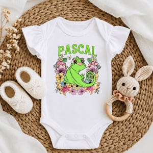 May include: A white baby bodysuit with ruffled sleeves featuring a graphic print of a green chameleon surrounded by colorful flowers and the text PASCAL in green letters. The garment includes a snap closure at the bottom for easy access.