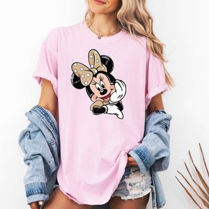 May include: A pale pink t-shirt with a Minnie Mouse cartoon graphic. The character has a polka dot bow, black ears, and a smiling face. The shirt has short sleeves and a loose fit.