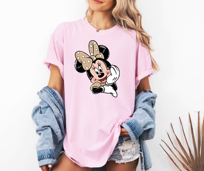 May include: A pale pink t-shirt with a Minnie Mouse cartoon graphic. The character has a polka dot bow, black ears, and a smiling face. The shirt has short sleeves and a loose fit.