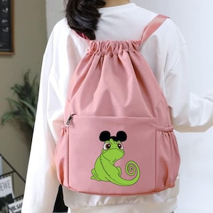 May include: A pink drawstring backpack with a front pocket and black zipper. The backpack features a green cartoon lizard wearing black Mickey Mouse ears. The straps are pink.