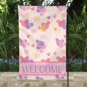 May include: A garden flag with a light pink background featuring a pattern of purple, pink, and yellow Mickey Mouse-shaped balloons and fireworks. The flag has a pink banner at the bottom with the word "WELCOME" in purple.