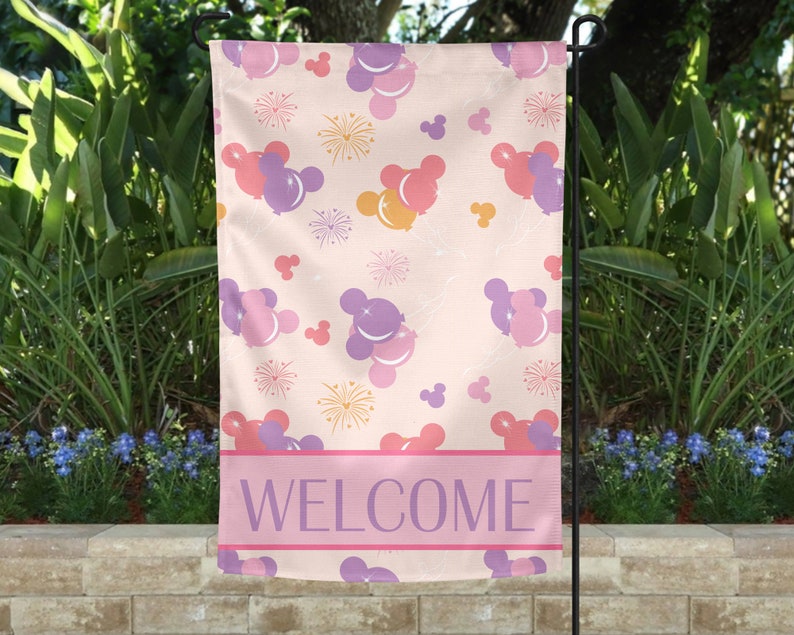 May include: A garden flag with a light pink background featuring a pattern of purple, pink, and yellow Mickey Mouse-shaped balloons and fireworks. The flag has a pink banner at the bottom with the word "WELCOME" in purple.
