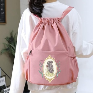 May include: A pink drawstring backpack with a front pocket and side pockets. The backpack features a framed illustration of a woman with long hair holding a rose. The straps are pink.