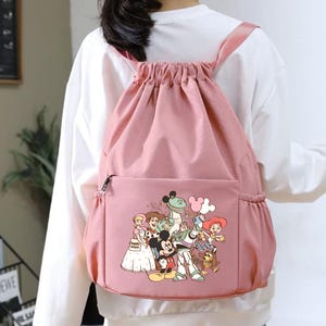May include: A pink drawstring backpack with a front pocket and a zipper closure. The backpack features a colorful graphic of cartoon characters. The straps are pink and the bag is made of a soft material.