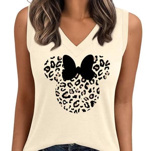 May include: Cream-colored sleeveless top with a V-neck. The front features a black bow and a leopard print design in the shape of a mouse head. The top is paired with blue jeans.