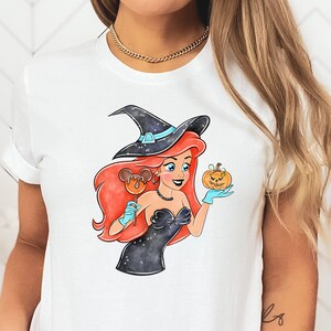May include: White t-shirt featuring a Halloween-themed illustration. The graphic depicts a cartoon character with red hair, wearing a witch hat and black dress, holding a pumpkin and a Mickey Mouse-shaped lollipop. The character is wearing blue gloves.