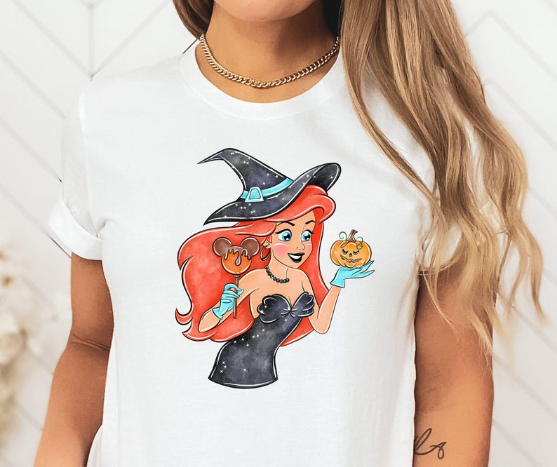 May include: White t-shirt featuring a Halloween-themed illustration. The graphic depicts a cartoon character with red hair, wearing a witch hat and black dress, holding a pumpkin and a Mickey Mouse-shaped lollipop. The character is wearing blue gloves.