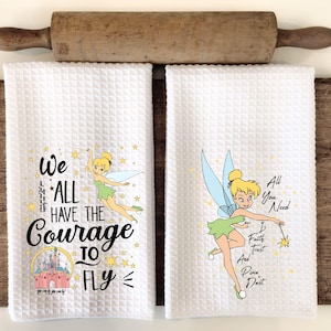 May include: Two white waffle-weave kitchen towels with Tinkerbell illustrations and inspirational quotes. One towel reads "We All Have The Courage To Fly" with a castle graphic. The other says "All You Need Is Faith, Trust, and Pixie Dust."