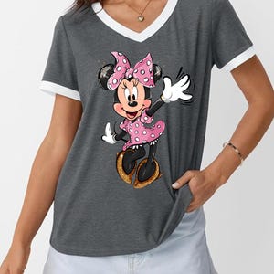 K&ouml;nnte beinhalten: Graues V-Ausschnitt-T-Shirt mit wei&szlig;em Besatz und Minnie Mouse-Grafik. Minnie tr&auml;gt ein rosa gepunktetes Kleid und eine Schleife mit goldenen Glitzerschuhen. Das T-Shirt hat einen l&auml;ssigen Stil.