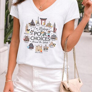 May include: A white V-neck t-shirt featuring a colorful graphic print of global landmarks, cartoon characters, and drinks arranged in a circle. The text reads "I'm Making POOR CHOICES Around The World" with labels for countries like France, Japan, and Mexico.