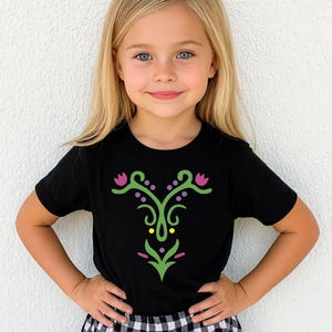 May include: Black short-sleeved t-shirt with a decorative floral design in green, purple, and yellow. The design is centered on the front of the shirt. The shirt is worn by a young child.