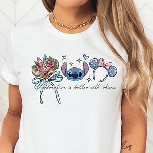 May include: White t-shirt featuring a colorful graphic of Stitch, Mickey Mouse ears, and a ukulele. The design includes the phrase "Adventure is better with 'ohana" in a script font. The design is in shades of blue, pink, and brown.